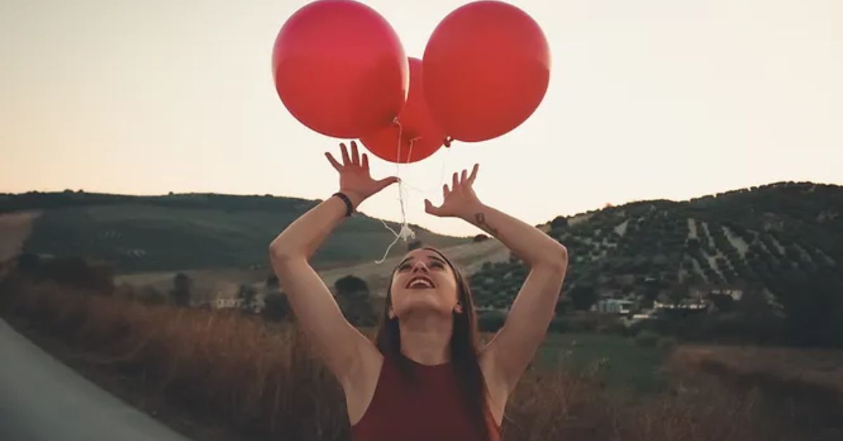 A girl looking at 3 red balloons above her head as she walks on trail with the mountains behind her.