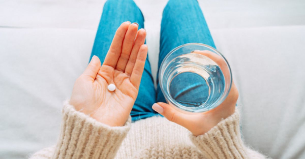 Woman in blue jeans and a sweater holding a glass of water and a pill